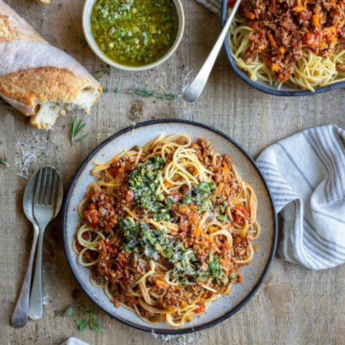 Vegane Linsen Bolognese schoen angerichtet. Mit Spaghetti auf einem weißen Teller mit blauem Rand, dazu Baguette.