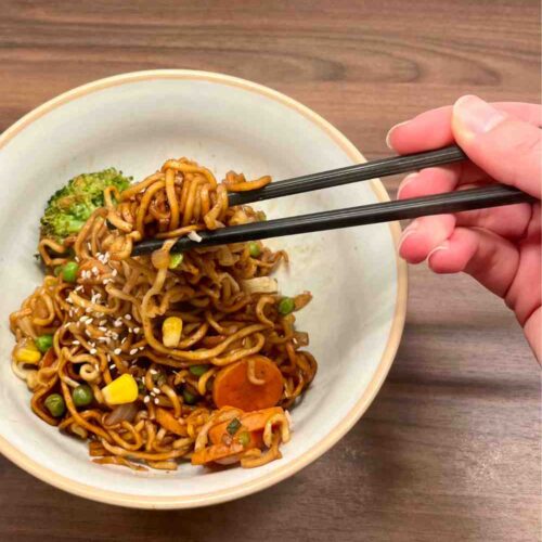 Asiatisch gebratene Nudeln mit Brokkoli, Karotten, Erbsen und Mais, mit Sesam bestreut, die mit Staebchen gegessen werden.